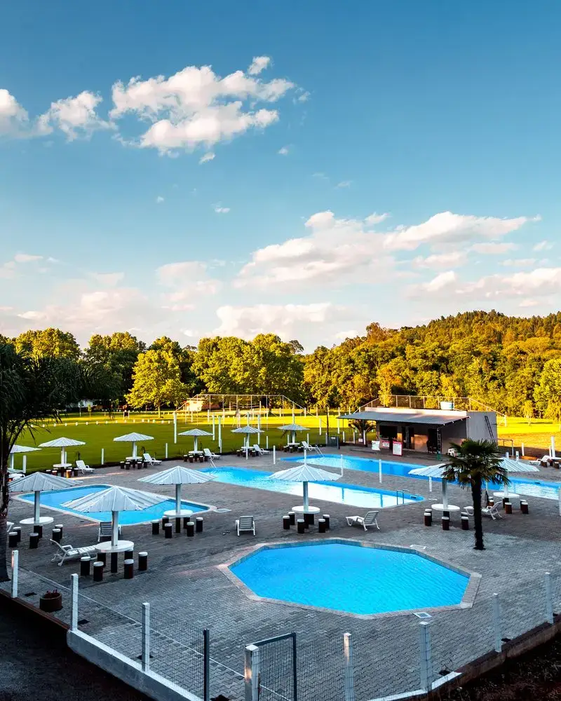 Vista do alto das piscinas, com guarda-sóis, gramado e morros ao fundo